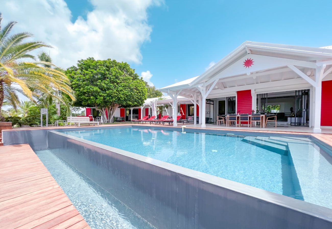 Vista de la piscina infinita, la amplia terraza y la parte delantera de la casa con mesa exterior y tumbonas rojas en una villa de alta gama en las An