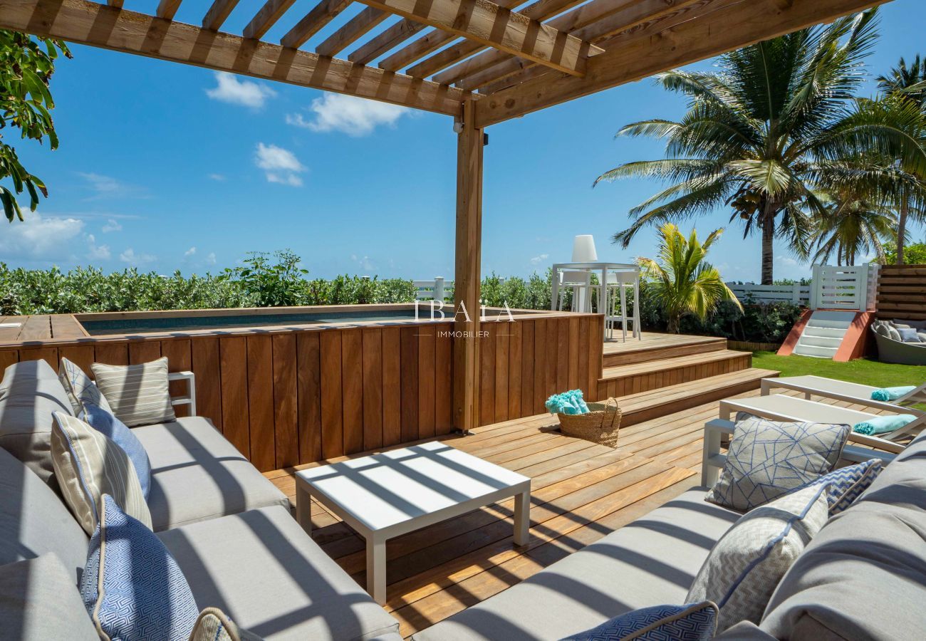 Vue du salon de jardin à côté de la piscine surélevée avec coffrage en bois, sous un patio ombragé en bord de mer